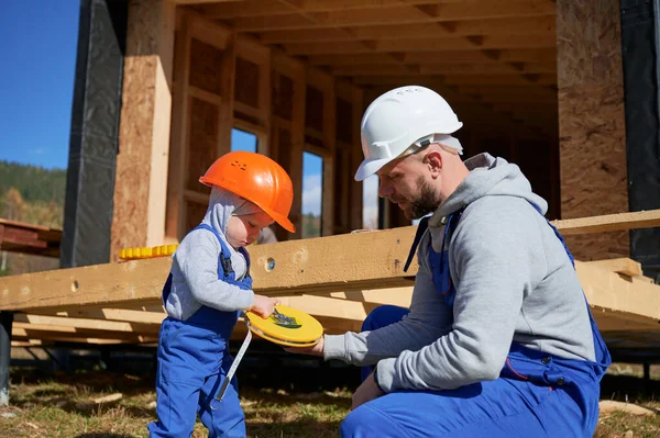 Father with toddler son building wooden frame house in Scandinavian style barnhouse. Boy helping his daddy, playing with tape measure on construction site on sunny day. Carpentry and family concept.
