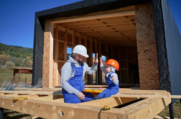 Küçük oğlu olan baba ahşap çerçeve inşa ediyor. Erkek müteahhit, şantiyedeki çocuğa güneşli bir günde kask ve mavi tulum giyerek beşlik çakıyor. Marangozluk ve aile kavramı.