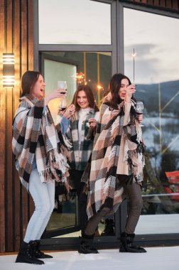 Young women enjoying winter weekends on terrace of contemporary barnhouse. Four girls in plaids drinking wine and celebrating with sparklers in the evening.