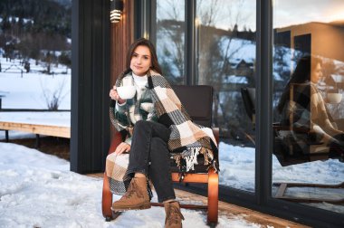 Young woman resting on terrace of modern barn house in the mountains. Happy female tourist sitting in chair, holding cup of tea, enjoying in new cottage in winter.