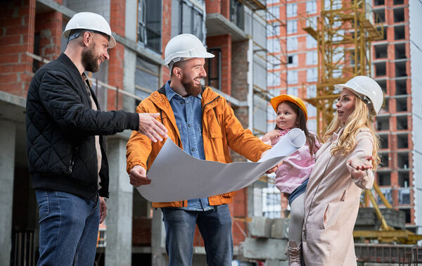 Happy family with child discussing architectural building plan with specialist at construction site. Parents with child and male builder standing outdoors near building under construction.