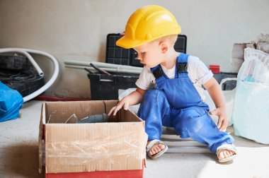 Güvenlik kaskı takmış şirin bir çocuk karton kutunun yanında ev yenileme ve onarım malzemeleri ile oturuyor. Çocuk inşaat işçisi karton kutudaki malzemelere bakıyor..