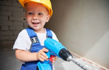 Şirin bebek inşaat işçisi elinde oyuncak elektrikli matkapla yan yana bakıp gülümsüyor. Tamir için elektrikli aletleri olan çocuk güvenlik kaskı takıyor ve iş tulumu giyiyor. Ev yenileme konsepti.