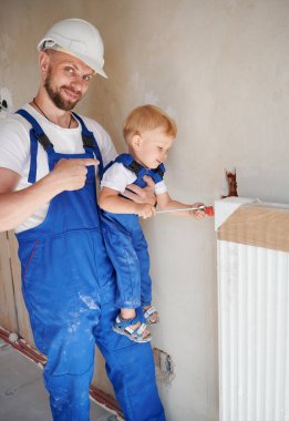 İş yerindeki tesisatçı bebek tulumunu gösterip gülümserken çocuk da tamirat altındaki daireye ısıtma radyatörü yerleştiriyor. Gururlu baba, küçük oğlunu evde inşaat işçisi olarak tutuyor..
