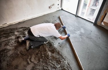 Erkek inşaat işçisi yere çimento karışımı bir şey döşemiş. Adam dairede zemini tararken düz kenarlı düzleştiriyor ve düzleştiriyor.