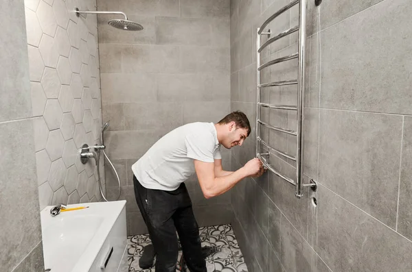 Man plumber hanging electric towel rack dryer on the wall with ceramic tile in bathroom. Male worker installing bath towel heater in apartment under renovation. Concept of home renovation.
