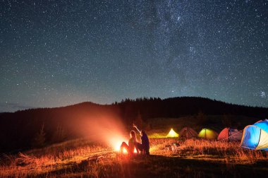 Yıldızlı gökyüzünün altındaki dağlarda gece kampı. Yanan kamp ateşinin yanında çimlerin üzerinde oturan iki kişinin silueti. Yıldızlarla dolu güzel gökyüzünü gösteren adam. Turizm ve sağlıklı yaşam kavramı.