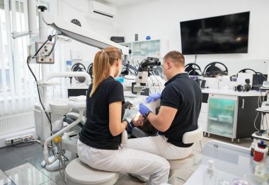 Konsantre olmuş dişçilerin sandalyeye oturup dişlerini düzelttiği arka plan. Üniforma, maske ve eldiven giyen iki doktor çalışıyor, teftiş ediyor, inceleme yapıyor. Stomatoloji kavramı.