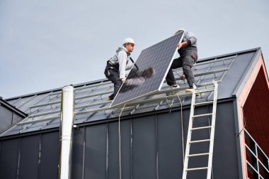 Kurucular evin çatısına güneş paneli kuruyorlar. Dışarıda fotovoltaik güneş modülü taşıyan miğferli işçiler. Alternatif ve yenilenebilir enerji kavramı.