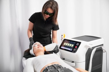 Güvenlik gözlüklü kozmetolog yüz bakımı yaparken diyot lazer cihazı kullanıyor. Kozmetik ekipmanlarının yanında yatan ve güzellik salonunda yüz gençleştirme tedavisi gören bir kadın..