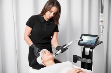 Hasta için yüz gerdirme ameliyatı yaparken radyoaktivite mikronezi cihazı kullanan kadın kozmetikçi. Kozmetik ekipmanlarının yanında yatan ve cilt bakımı gören kadın..