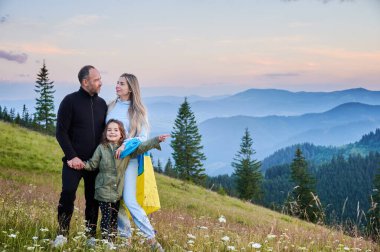 Lovely Ukrainian family resting in mountains. Male adult and his wife standing with their child that pointing at something. Carpathian mountains landscape. clipart