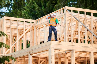 Carpenter building wooden frame two-story house. Bearded man wearing protective goggles carrying chainsaw on his shoulder, dressed in protective helmet, coveralls, and orange vest. clipart