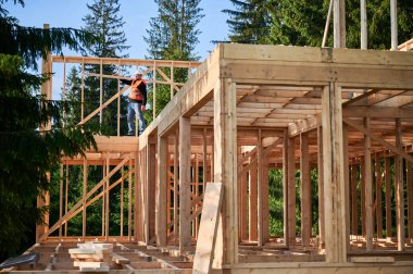 Marangoz ormanın yakınında ahşap çerçeveli ev inşa ediyor. Adam omzunda büyük bir kalas tutuyor ve iş kıyafetleri ve miğfer giyiyor. Çevreye duyarlı modern yapı kavramı.