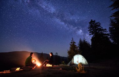 Dağlarda gece kamp gezisi, parıldayan yıldızların altında güneşlenme. Erkek ve kadın, maceraperest gezginler, şenlik ateşinin ve ormanın ortasına kurulmuş aydınlık çadırlarının yanında huzur bulurlar..