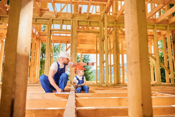 Father with toddler son constructing wooden frame house. Male builder showing to kid construction site, wearing helmets and blue overalls on sunny day. Carpentry and family concept.