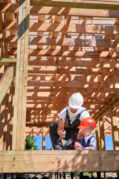 Father with toddler son building wooden frame house. Man instructing his son on the art of hammering nails on construction site, wearing helmets and overalls on sunny day. Carpentry and family concept