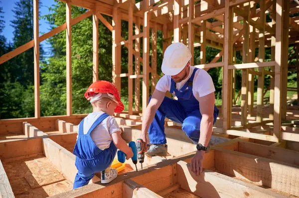 Father with toddler son building wooden frame house. Worker teaching his son how to use screwdriver on construction site, wearing helmets and blue overalls on sunny day. Carpentry and family concept.