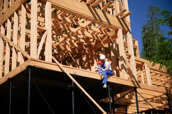 Father with toddler son building wooden frame house. Male builder sitting on the edge of the balcony at construction site, wearing helmets and blue overalls on sunny day. Carpentry and family concept.