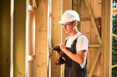 Marangoz ahşap çerçeveli ev inşa ediyor. Gözlüklü bir adam tornavidayla çalışıyor, iş tulumu ve kask giyiyor. Modern çevre dostu yapı kavramı.