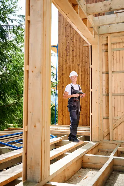 Portrait of carpenter constructing wooden framed house. Man worker cladding facade of house, using screwdriver. Concept of modern eco-friendly construction.