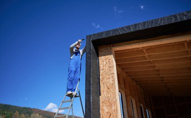 Erkek müteahhit, arka planda mavi gökyüzü, gelecekteki kulübenin ön cephesi olarak kullanılan siyah kıvrımlı demir çarşaf kuruyor. Ahşap çerçeve inşa eden bir işçi. Marangozluk ve inşaat konsepti.