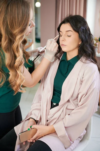Female makeup artist applying eyeshadow on client eyelid with cosmetic brush. Close up of young woman keeping eyes closed and smiling while stylist doing professional eye makeup.