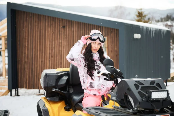Portrait of beautiful woman posing on offroad four-wheeler ATV with wooden house on background. Concept of active leisure and winter activities.