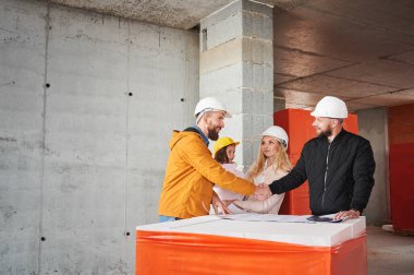 Çocuklu bir aile, inşaat işçisiyle yeni dairenin mimari planını tartışıyor, inşaat halindeki apartmanın içinde duruyor. Adam inşaatçıyla el sıkışıyor..