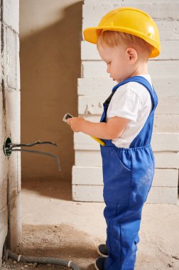 Duvara elektrik kablosu ve soket yerleştirirken tel kesici kullanan sevimli bir çocuk inşaat işçisi. Güvenlik kasklı bir çocuk, tadilat altındaki dairede elektrik kabloları monte ediyor..