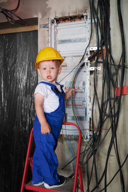 İnşaat kaskı takan, elinde elektrik kablosu tutan ve dairede elektrik kontrol panelini tamir ederken gülümseyen sevimli küçük çocuk. Elektrik santralinin yanındaki merdivende duran çocuk elektrikçi..
