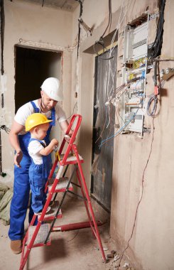 Bebek elektrikçi merdivende duruyor ve evde babasıyla birlikte telefon santrali kurarken pense aletini tutuyor. Güvenlik kasklı bir adam ve çocuk, tadilat altındaki apartmanda elektrik kabloları yerleştiriyorlar..