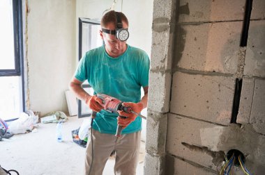 Erkek inşaat işçisi, yenilenmekte olan dairede elektrikli matkapla tuğla duvar sondajı yapıyor. Evde profesyonel matkap kullanırken fener ve iş eldiveni takan bir adam..