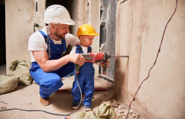 Bir adam ve küçük çocuk inşaat işçileri tadilat altındaki dairede elektrikli matkapla duvar kazıyorlar. Evde profesyonel sondaj makinesi kullanan kasklı erkek ve çocuk..