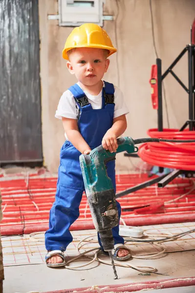 Güvenlik kaskı takmış elinde elektrikli matkap olan ve kameraya bakan bir çocuk. Profesyonel sondaj aletli çocuk inşaat işçisi apartmanda tadilat altında duruyor..