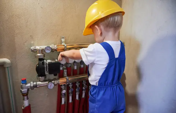 Baby boy checking plumbing installation with pipes and thermometer gauge. Kid plumber in workwear working with plumbing pipe system in apartment under renovation.