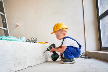 İş yerindeki sevimli çocuk, tadilat altındaki dairede elektrikli inşaat aletleri kullanıyor. Güvenlik kasklı şirin çocuk tadilat sırasında evde elektrikli matkapla çalışıyor..
