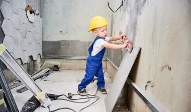 İnşaat miğferi takmış çocuk, duvarın yüzeyini ruh seviyesiyle kontrol ediyor. İş yerindeki bir çocuk, ev tadilatında çalışırken aynı seviyedeki aletleri kullanıyor.