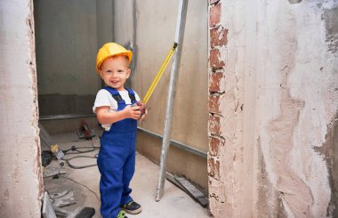 Mutlu inşaat işçisi elinde şerit tutarken ve dairede tadilat sırasında gülümserken. Güvenlik kaskı takan ve iş tulumu giyen neşeli bir çocuk..