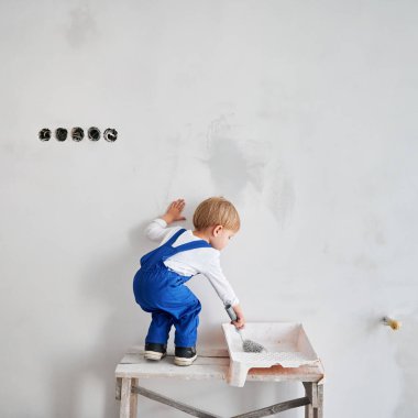 Apartman dairesine beyaz duvar boyayan sevimli küçük bir çocuğun arka manzarası. İş yerindeki çocuk ahşap masanın üzerinde duruyor ve evde tadilat yaparken boya silindiri kullanıyor..