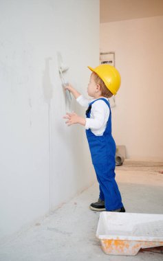 Full length of child construction worker painting wall in apartment under renovation. Kid in safety helmet and work overalls using paint roller while working on home renovation.