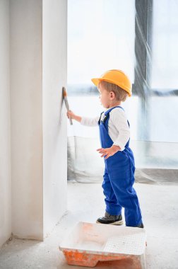 Çocuk inşaat işçisi dairede boya fırçasıyla duvar boyuyor. Güvenlik kaskı takan ve evde tadilat sırasında boya fırçası kullanan çocuk..