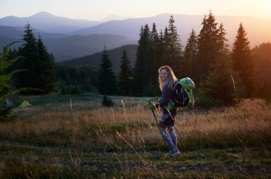 Kadın turist gün batımında dışarıda yürüyüş yapıyor. Dağlarda gezen sportif, pozitif bir kadın. Sırt çantalı genç, sportif bir kadın yürüyüş sopası kullanıyor, yürüyor, gülümsüyor. Doğayla uyum kavramı.