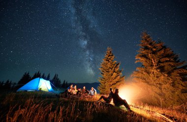 Night camping in mountains under starry sky. Group of people tourists having a rest near campsite, burning campfire and illuminated tent. Concept of tourism, hiking and adventure.