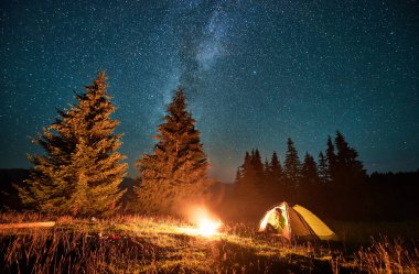 Yıldızlı gökyüzü ve Samanyolu 'nun altındaki dağlarda gece kampı. Kamp alanındaki çadırda oturan dişi turist manzaraya ve doğaya hayranlık duyuyor, ateş yakıyor. Seyahat ve yürüyüş kavramı.