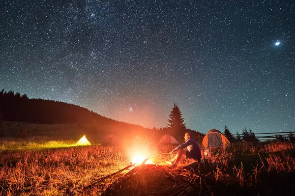 Night camping in mountains under starry sky. Man sitting on grass near fire, holding stick. Male hiking, traveling, admiring fire, landscape, resting, enjoying. Concept of harmony with nature.