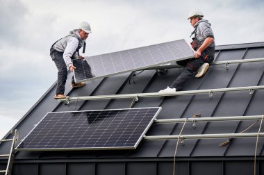 Evin çatısına güneş paneli inşa eden mühendisler. Dışarıda fotovoltaik güneş modülü taşıyan miğferli işçiler. Alternatif ve yenilenebilir enerji kavramı.