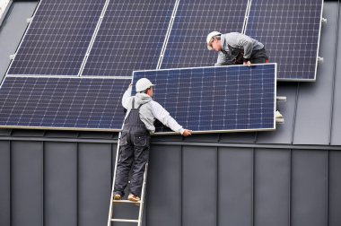 Mühendisler evin çatısına fotovoltaik güneş panelleri kurarken kabloları bağlıyorlar. PV güneş modüllerini çatıya monte eden işçiler. Alternatif ve yenilenebilir enerji kavramı.