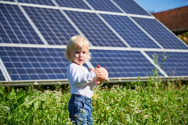Little cute boy saving money in piggy bank. Small kid learning about saving money for future. Blond child holding piggy bank on background of solar panels at sunny day.