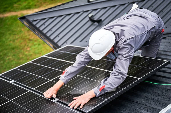 Man builder mounting photovoltaic solar panels on roof of house. Engineer in helmet installing solar module system with help of hex key. Concept of alternative, renewable energy.